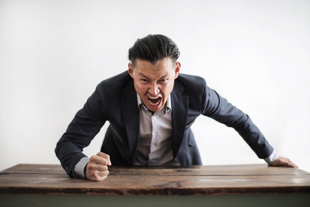 Expressive angry boss in formal suit looking at camera and screaming with madness while hitting desk with fist